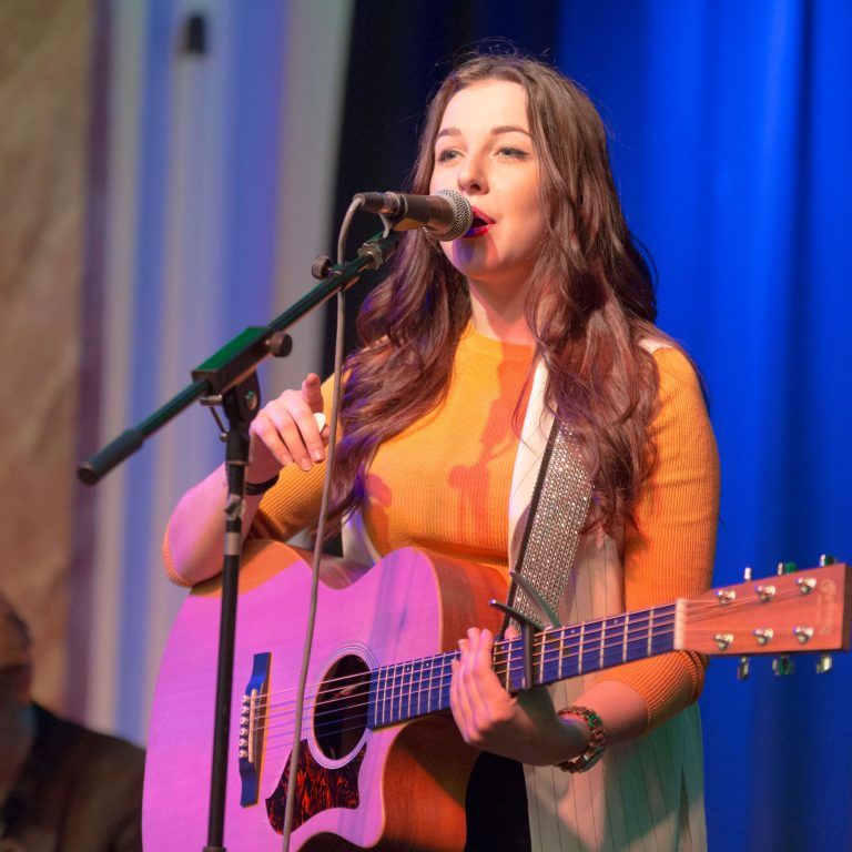 Young woman on stage singing into a microphone and playing an acoustic guitar