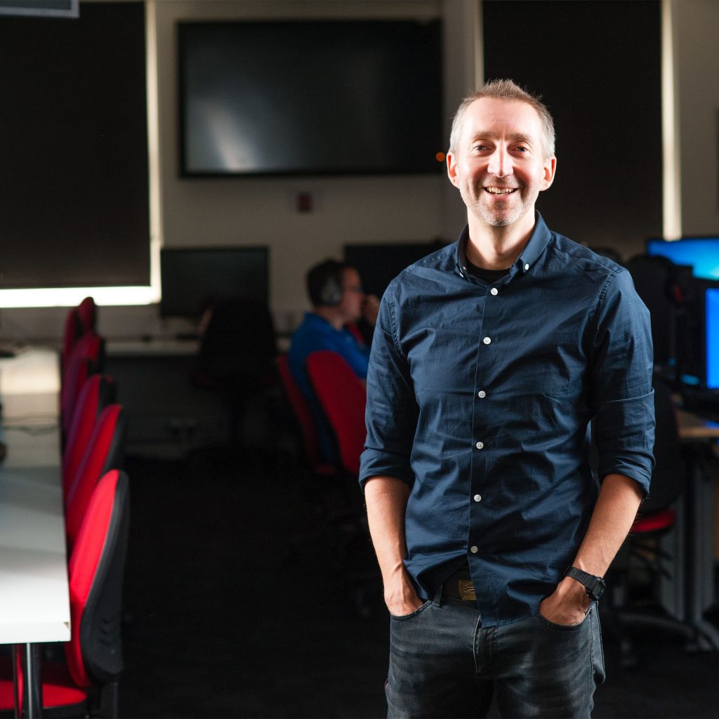 Richard Cook, Postgraduate Assistant in Games Design looking at camera with seminar room in background