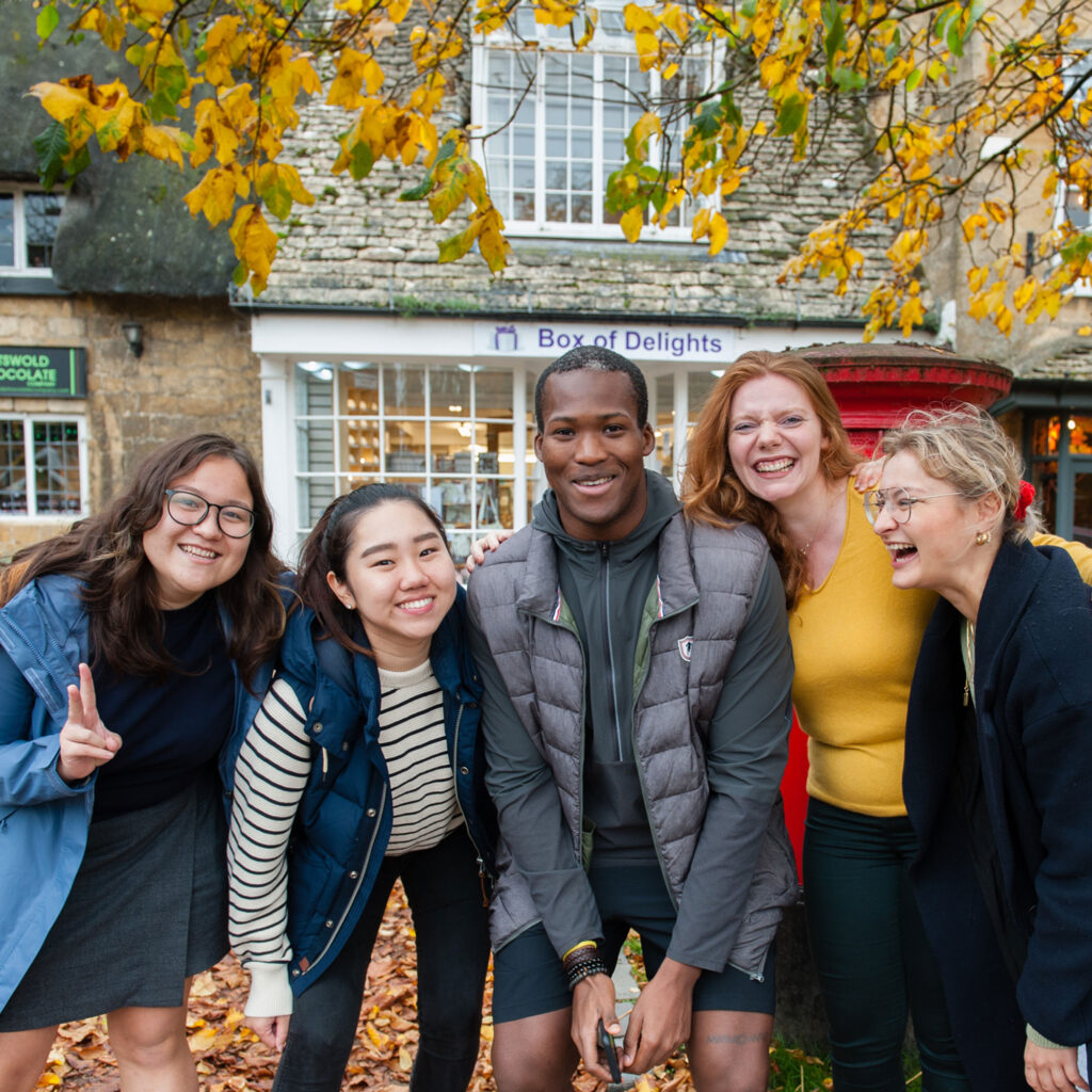 Students laughing in front of shops in a Cotswold town with autumn leaves in the background.