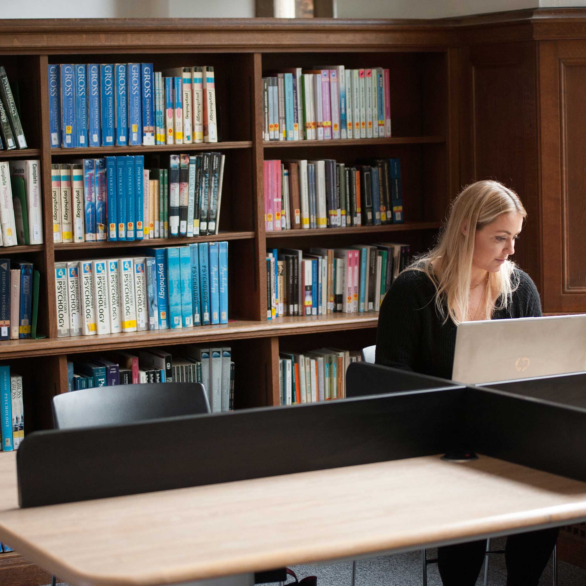 Postgraduate Psychology student working at a laptop on a desk in a library, with Psychology textbooks on the shelf behind her.