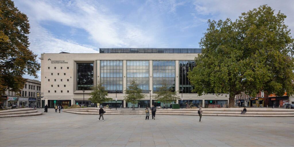 A front external view of the City Campus building, which features large vertical glass windows and “University of Gloucestershire” signage set on the top left side of the building. A few people walk across the paved public area in the foreground, mature trees frame both sides and a blue sky above with some wispy clouds.