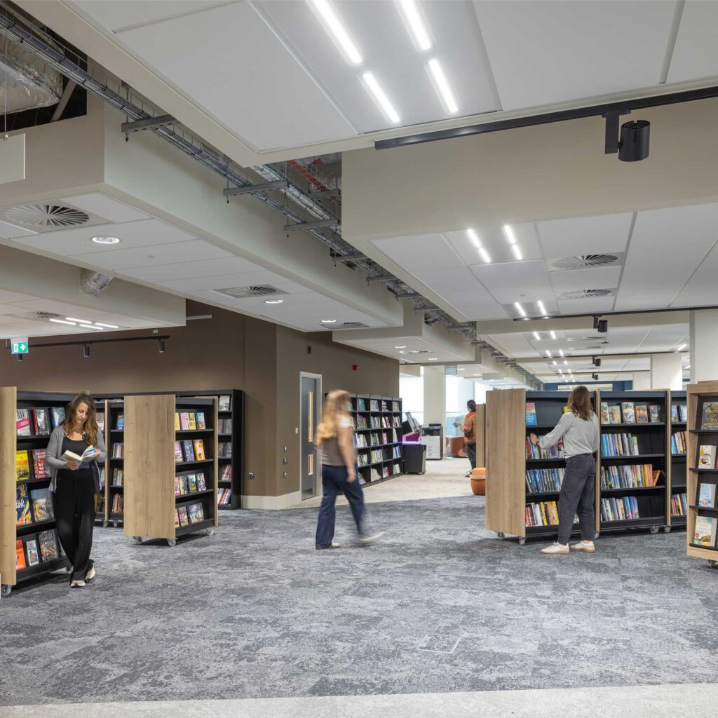 Interior image of the library at City Campus with rows of wooden bookshelves filled with books and grey carpeted floor. Several people browse the shelves with one person standing reading a book near the foreground.