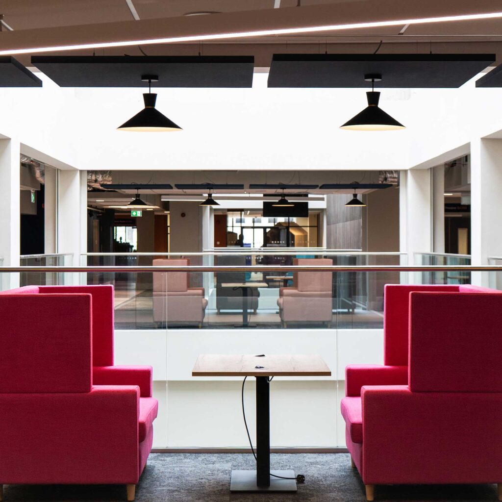 An agile working area with two bright pink high-backed armchairs arranged around a small table beside a glass balustrade, overlooking an open-plan atrium. There are hanging black pendant lights and seating areas in the background.