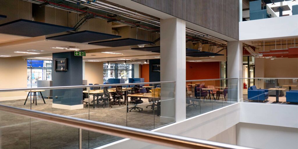 Open-plan agile working space at City Campus. Workstations with office chairs and desks are arranged on the left, and seating with blue and purple chairs are visible on the right.