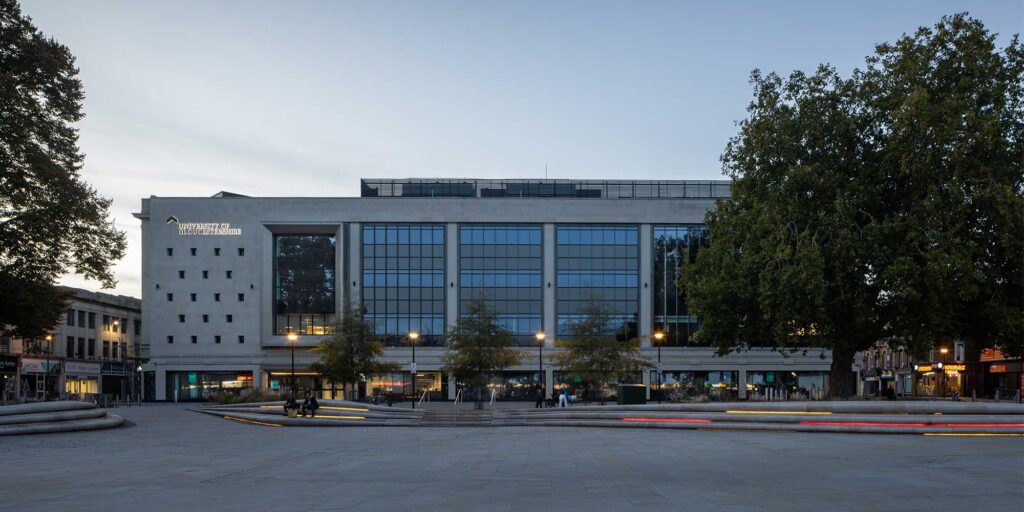 A front external view of the City Campus building during the evening. Mature trees frame both sides of the building and coloured strip lights display on steps in front.