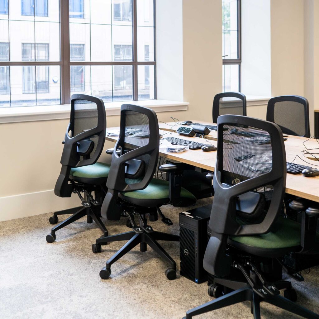 Large office desk with keyboards and mice surrounded by five office chairs. A window in the background looking out on other buildings.